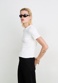 Person with short blond hair wearing black sunglasses, white short-sleeve shirt, black pants, and a cherry tattoo on their left arm.