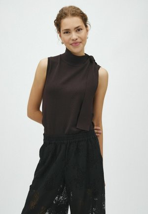 Vrouw met krullend haar, gekleed in een zwarte mouwloze top met nekstrik en zwarte kanten wijde broek, staand met één hand achter haar rug.