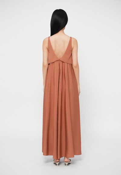 Γυναίκα με μακριά μαύρα μαλλιά, ντυμένη με μια αμάνικη, χάλκινη μάξι φόρεμα με χαμηλή πλάτη και μπεζ τακούνια, στέκεται μπροστά σε έναν απλό φόντο.