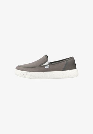 Zapato gris tipo slip-on con suela de goma blanca con motas y pequeña etiqueta de tela en el lateral sobre fondo blanco.