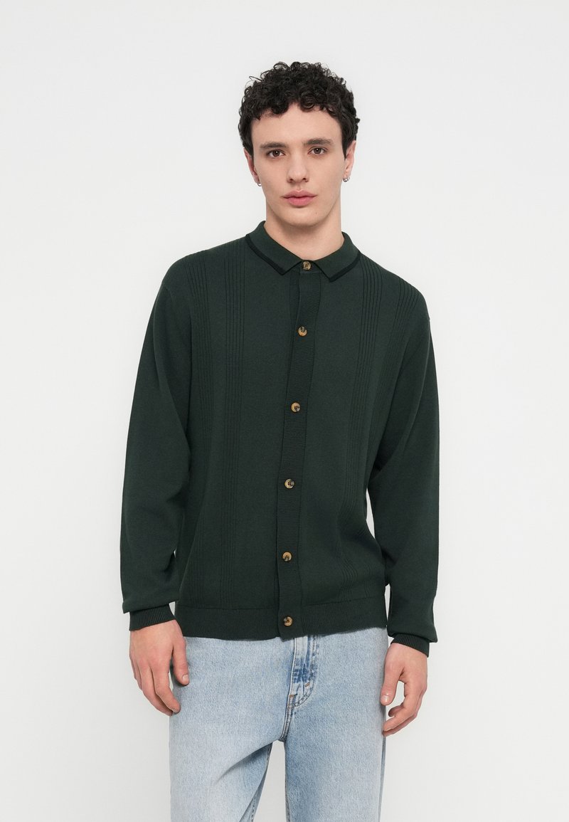 Σκούρα πράσινη ζακέτα με κουμπιά και γιακά, με ανάγλυφη υφή και έξι χρυσά κουμπιά. Χαλαρή εφαρμογή με μακριά μανίκια.