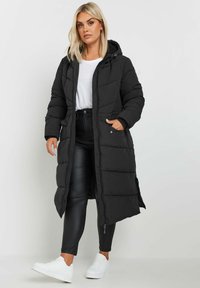 Zwarte pufferjas met capuchon, lange lengte, gewatteerde textuur, voorzakken en een ritssluiting. G gedragen met zwarte broek en witte sneakers.