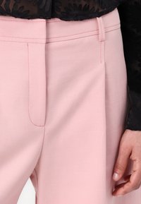 Světlé růžové kalhoty mají texturovanou látku, ploché přední kapsy a kontrastní červené prošívání podél švů.