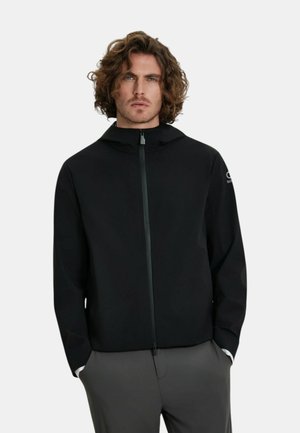 Uomo con capelli ricci che indossa una giacca nera con cappuccio e zip e pantaloni grigi, in piedi con le mani nelle tasche davanti a uno sfondo semplice.