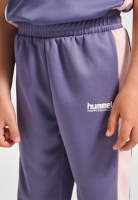Lila Jogginghosen mit elastischem Bund, mit einem pinken Seitenstreifen und weißem „hummel PERFORMANCE“-Logo am unteren Bein. Glatte Stoffstruktur.