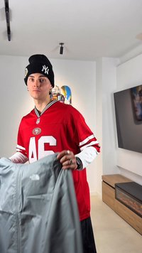 Mladá osoba v červenom drese San Francisco 49ers a čiernej čiapke drží sivú bundu North Face v modernom obývacej izbe.