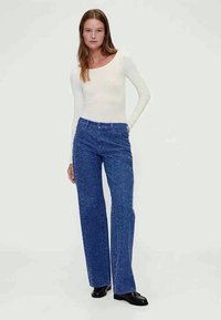 Blue denim wide-leg pants with a textured finish, high waist, and standard pockets, paired with a fitted long-sleeve white top.