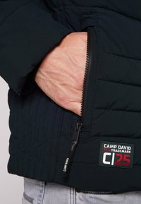 Giacca imbottita color navy con tasca con zip, dotata di una cerniera nera e un'etichetta del marchio. Tessuto testurizzato con dettagli di cucitura distintivi.