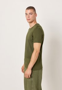 Olivno zelena bombažna majica z okrolim ovratnikom in kratkimi rokavi, nošena s svetlo zelenimi hlačami, prikazana na nevtralnem ozadju.