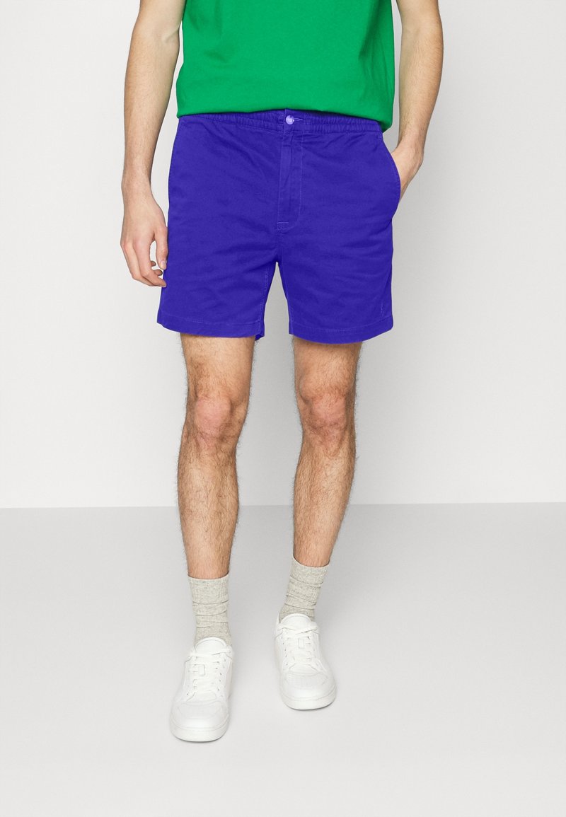 Homme portant un short bleu vif, une chemise verte, des chaussettes beiges et des baskets blanches, debout devant un fond clair uni.
