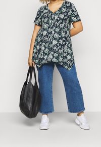 Túnica estampada de flores en navy, con mangas cortas y escote en V. Combinada con vaqueros de pierna ancha azules y un bolso de cuero negro. Zapatillas blancas.