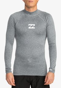 Rash guard de manga larga gris con un diseño ajustado, hecho de una tela suave y elástica. Presenta un logo blanco en el pecho y orificios para los pulgares.
