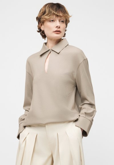 Γυναίκα με κοντά μαλλιά που φοράει μπεζ μπλούζα μακρυμάνικη με άνοιγμα στο λαιμό και λευκό ψηλόμεσο πλισέ παντελόνι, με τα χέρια στις τσέπες.