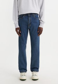 Person som bär blå rak ben-jeans, vita sneakers och en ljusgrå långärmad skjorta mot en helt vit bakgrund.