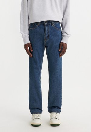 Person wearing blue straight-leg jeans, white sneakers, and a light gray long-sleeve shirt against a plain white background.