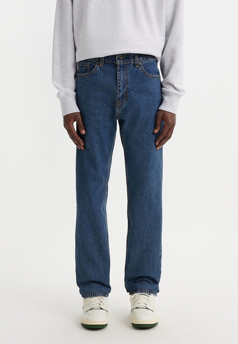 Person som bär blå rak ben-jeans, vita sneakers och en ljusgrå långärmad skjorta mot en helt vit bakgrund.