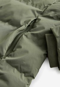 Jachetă umplută de culoare verde măsliniu, cu un buzunar cu fermoar, textură netedă și manșete côtate. Materialul pare ușor, dar totodată izolat.