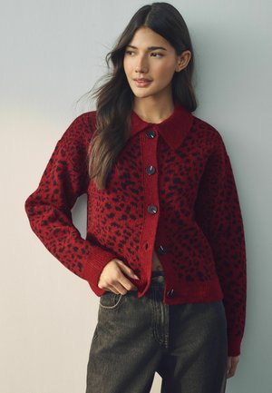 Red knitted cardigan with black leopard spots, featuring a pointed collar and black buttons, paired with dark denim jeans.