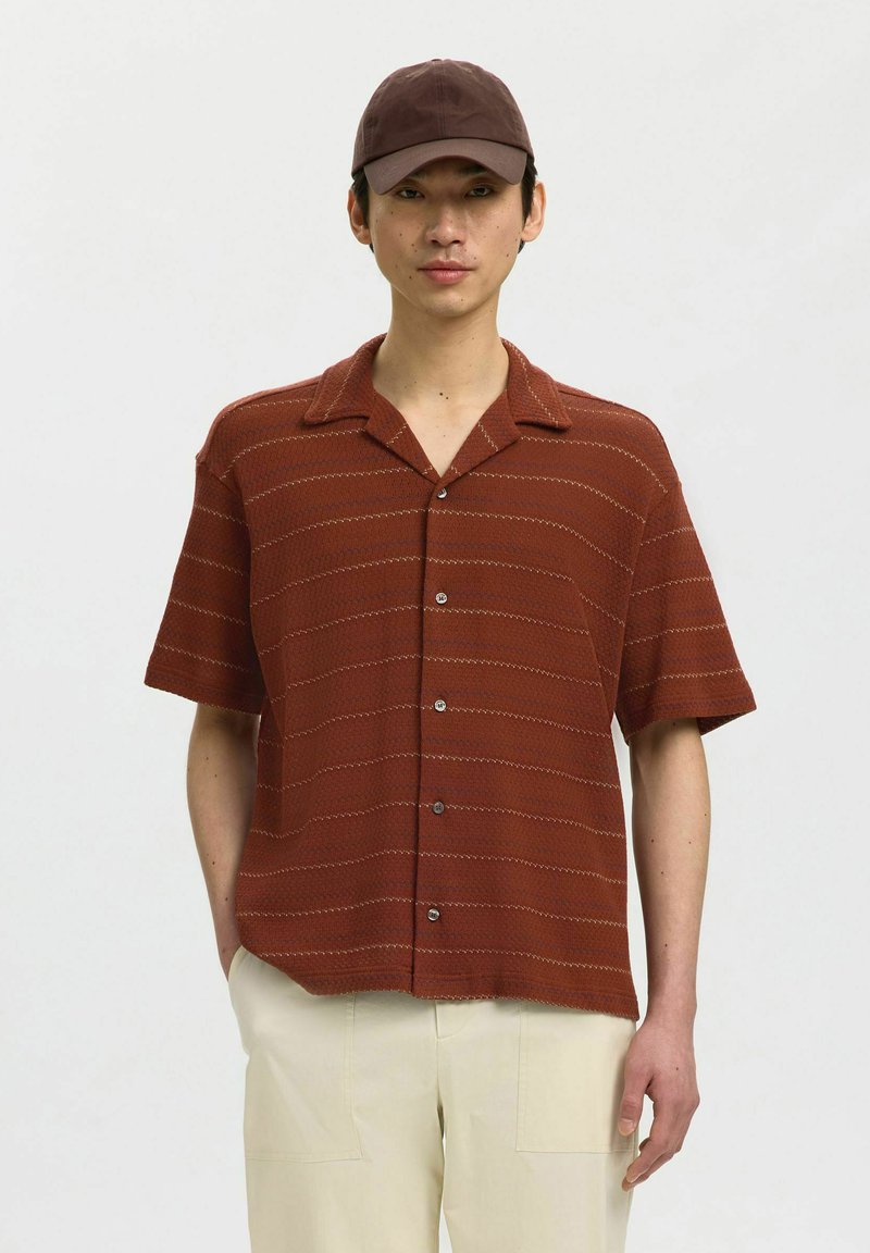 Jeune homme portant une chemise marron à manches courtes boutonnée avec des rayures subtiles, un pantalon beige et une casquette marron foncé sur un fond uni.