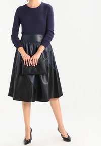 Vrouw in een marineblauw geribbeld sweater en zwarte leren midi-jurk, met een zwarte clutch en zwarte hoge hakken.