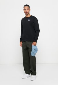 Man standing against white background wearing black sweatshirt, dark green pants, white sneakers, holding light blue cap in right hand.