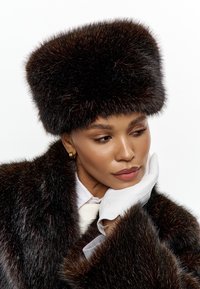 Faux furhatt med fyrkantig topp i mörkbrunt, med en mjuk textur och en glänsande yta, kombinerad med en mörk pälsjacka och vita handskar.
