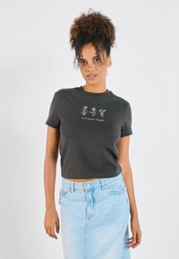 Mørk grå cropped t-skjorte med korte ermer, prydet med hvit blomsterbrodering og tekst foran, kombinert med et lyst denimskjørt.