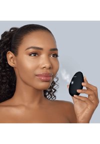 Un appareil portable noir avec une forme lisse et arrondie, émettant une fine brume. L'utilisateur a un teint de peau moyen et un maquillage naturel, tenant l'appareil près de son visage.