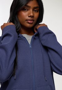 Giovane donna con capelli scuri che indossa una felpa con zip blu navy, tenendo il colletto con entrambe le mani su uno sfondo bianco.