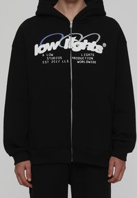 Hoodie noir zippé en tissu doux, arborant un grand motif graphique en texte blanc et bleu sur la poitrine et des poches latérales.