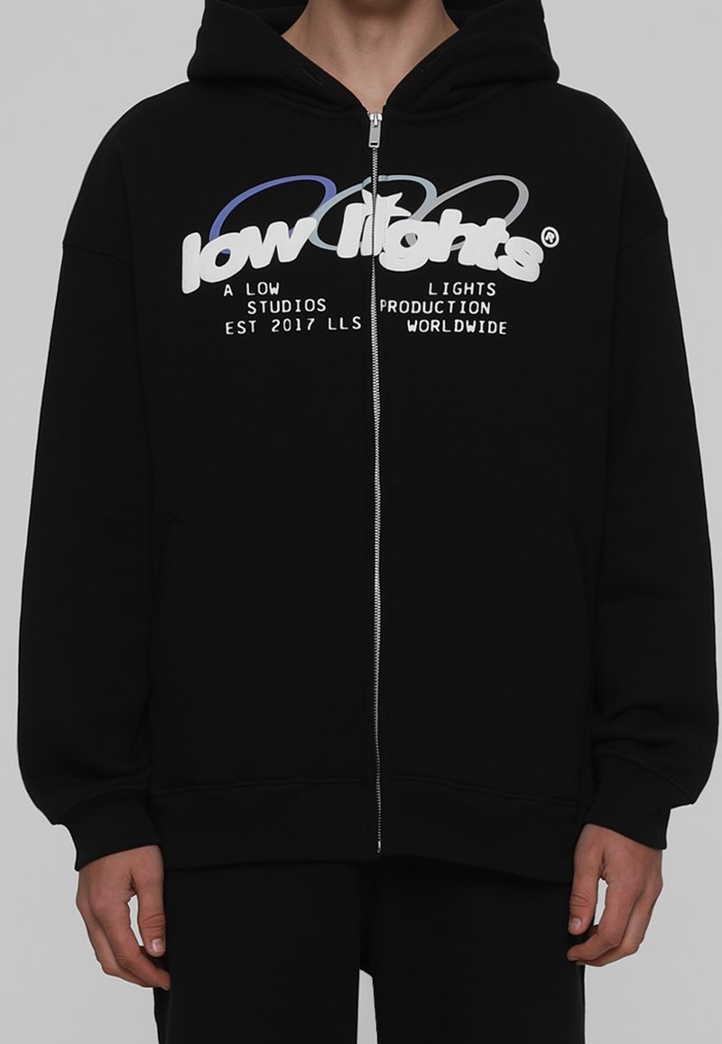 Hoodie noir zippé en tissu doux, arborant un grand motif graphique en texte blanc et bleu sur la poitrine et des poches latérales.