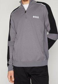 Mann trägt grauen und schwarzen Langarm-Pullover mit Viertelreißverschluss und BOSS-Logo auf der Brust, kombiniert mit schwarzen Hosen.