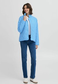 Jachetă matlasată de culoare albastru deschis, cu nasturi cu cap ștanțat, guler înalt și două buzunare, asortată cu blugi blue și adidași albi.
