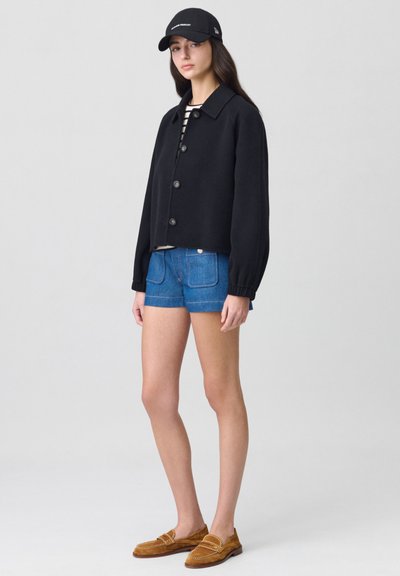 Veste noire courte avec des boutons, chemise rayée en dessous, short en denim bleu, et mocassins en suède brun. Portée avec une casquette noire.