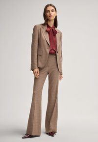 Completo marrone a quadri con pantaloni a zampa d'elefante e una blusa bordeaux con dettaglio a fiocco, accompagnato da scarpe lucide bordeaux con punta affusolata.