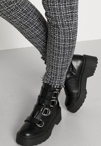Botas de tornozelo em couro preto com várias tiras de fivela de metal e sola grossa. Combinadas com leggings texturizadas em padrão preto e branco.