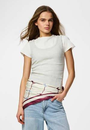 Noor naine pikkade pruunide juustega, kellel on seljas valge lühikeste varrukatega topp ja tank, sinised teksad ning vöökohale seotud mustriline sall.