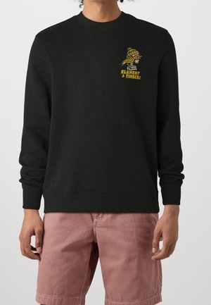 Schwarzer Sweatshirt mit langen Ärmeln und gelber Grafik eines Skeletts in Winterkleidung sowie dem Schriftzug "Element & Timber", kombiniert mit staubrosafarbenen Shorts.