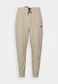 Beige mjukisbyxor av texturerat tyg med elastisk midja, dragsko och en liten svart logotyp på vänstra sidan.