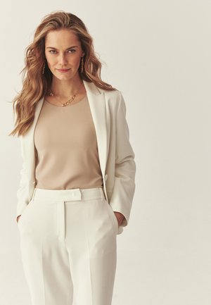 Žena s vlnitými blond vlasy, oblečená v krémovém saku a kalhotách s béžovým topem, zlatý náhrdelník a náušnice, ruce v kapse.