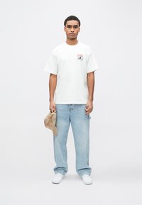 Λευκό κοντομάνικο μπλουζάκι με γραφική εκτύπωση στο στήθος, συνδυασμένο με ανοιχτό μπλε τζιν σε άνετη γραμμή και λευκά αθλητικά παπούτσια. Κρατάει ένα μπεζ καπέλο.