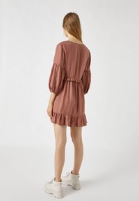 Rüschiges, kurzes braunes Kleid aus leichtem Stoff mit Puffärmeln und elastischem Bund, kombiniert mit klobigen weißen Stiefeln.