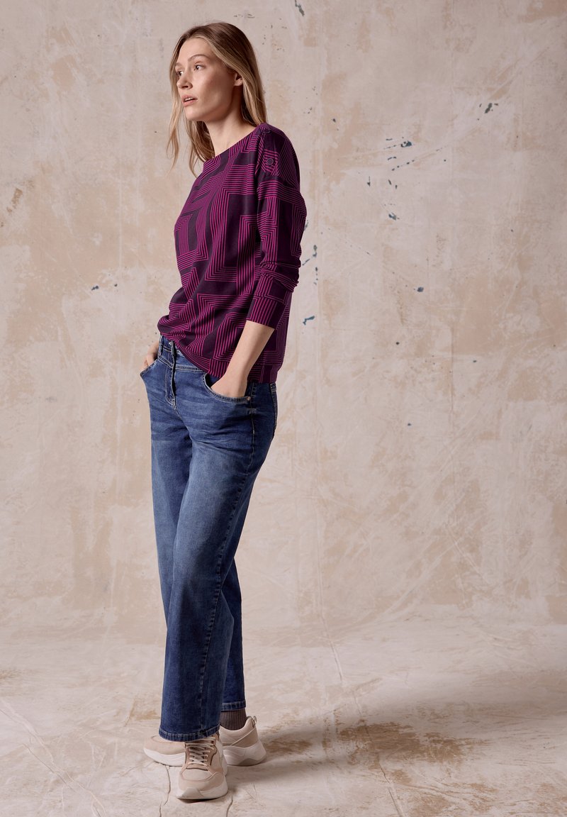 Top de manga larga morado con patrón geométrico, combinado con jeans de mezclilla azul y zapatillas beige. El modelo está de pie con las manos en los bolsillos.