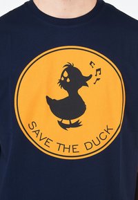 T-shirt in cotone blu navy con una grafica circolare gialla. Illustrazione di un'anatra nera all'interno con note musicali e il testo "SAVE THE DUCK" sotto.