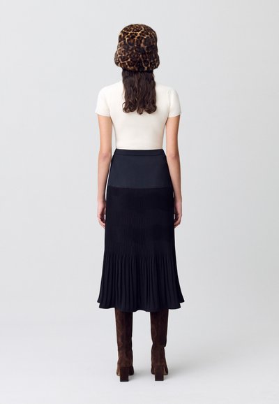 Falda maxi negra plisada con dobladillo acampanado, combinada con una blusa ceñida color crema y un sombrero de piel sintética con estampado de leopardo. Botas de ante marrón hasta la rodilla.