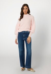 Helles rosa, flauschiges Sweatshirt mit Ballonärmeln, kombiniert mit hochtaillierten, dunkelblauen, weit geschnittenen Jeans und flachen goldenen Schuhen. Ein einfaches Gürtel-Detail ist enthalten.