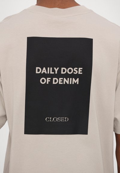 Beige bomull t-skjorte med et svart rektangulært grafisk motiv på baksiden, med hvit tekst som leser "DAILY DOSE OF DENIM" og "CLOSED."