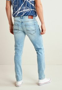 Jean en denim bleu clair à coupe slim, avec des poches arrière et des détails cousus. Associé à des baskets blanches et une chemise à motifs.