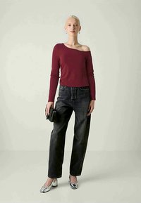 Top bordeaux de mangas longas com um design assimétrico, combinado com uns jeans pretos de cintura alta e sapatos de salto prateados. Pequena bolsa preta.