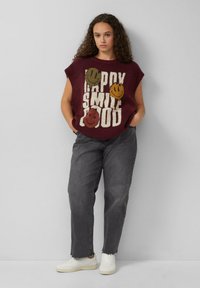 Frau mit lockigem Haar, die einen ärmellosen burgunderfarbenen Pullover mit Smiley-Gesichtern und weißem Text, graue Jeans und weiße Sneakers trägt.